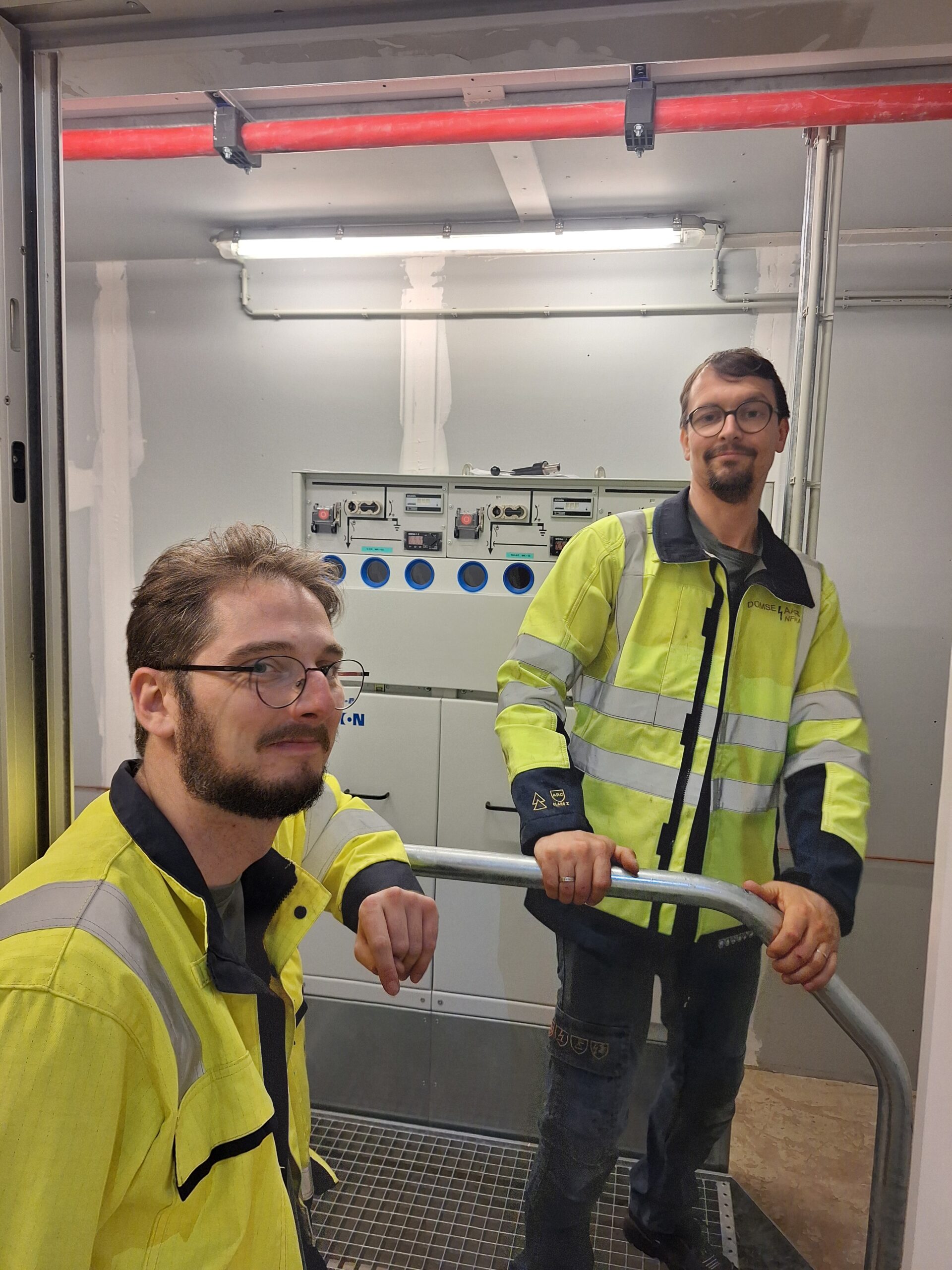 Twee medewerkers van NED in gele veiligheidsvesten staan voor een elektrisch bedieningspaneel in een industriële omgeving.