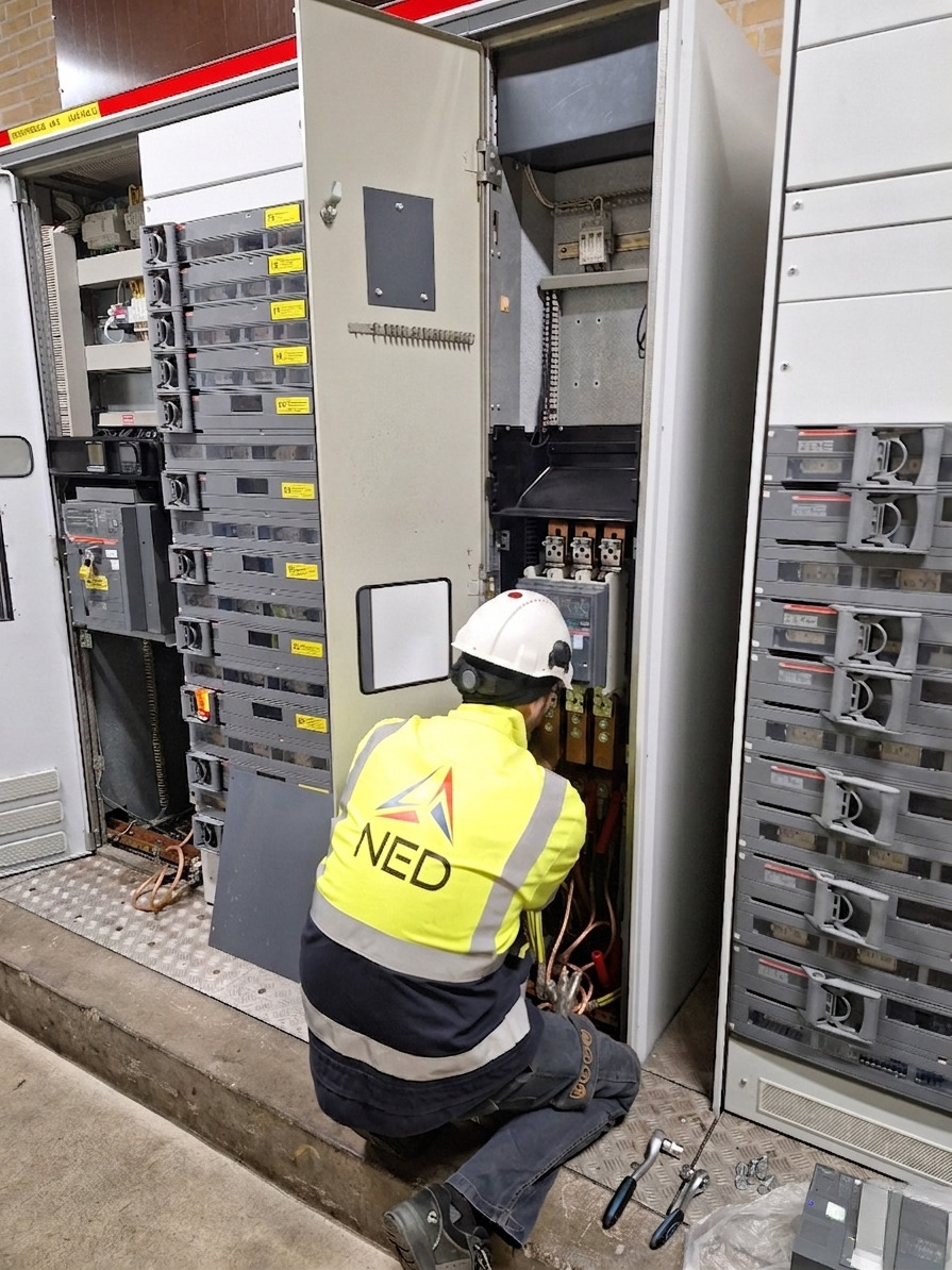 avico-project-NED Een arbeider met een veiligheidsvest en helm onderhoudt elektrische panelen met gereedschap dat naast hem op de grond ligt.