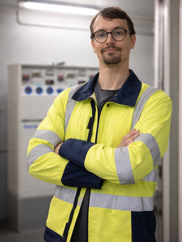 Marco van Domselaar met bril en veiligheidsvest, staand binnen met gekruiste armen voor industriële bedieningspanelen.