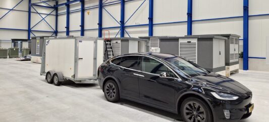 20231113_140530 Een zwarte Tesla Model X met aanhanger staat geparkeerd in een ruim industrieel pakhuis met blauwe balken en machines.