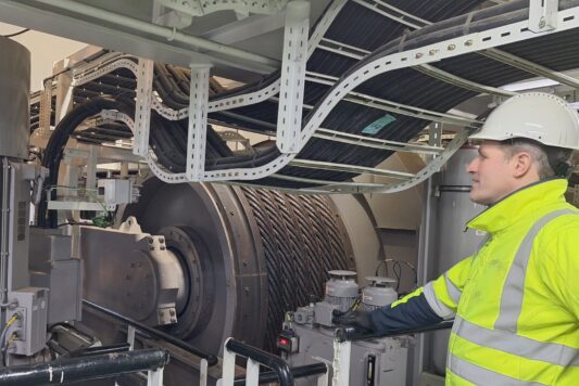 20250121_182357 Een werknemer met een helm op en een veiligheidsvest inspecteert grote industriële machines met meerdere kabels en metalen onderdelen.