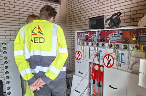 20250503_091800 Twee medewerkers van NED in zeer zichtbare kleding staan voor panelen met waarschuwingsborden voor elektrische apparatuur.