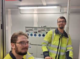 Twee medewerkers van NED in gele veiligheidsvesten staan voor een elektrisch bedieningspaneel in een industriële omgeving.