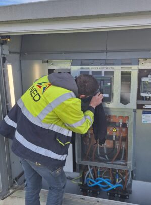 20260507_093343 Een arbeider in een high-visibility jas controleert apparatuur in een elektrisch controlepaneel buiten.