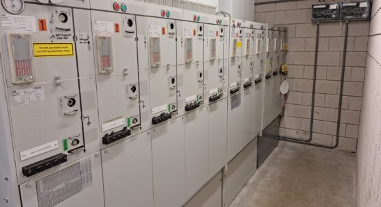 Een rij industriële elektrische regelpanelen met schakelaars, meters en waarschuwingslabels, geïnstalleerd langs een betonnen muur in een bijkeuken.
