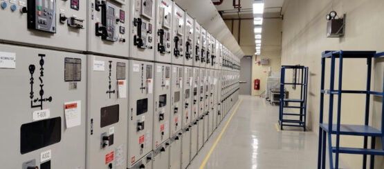 NED-web-cabinet-001 Een lange rij industriële elektrische regelpanelen omzoomt één muur van een schone, goed verlichte ruimte met rekken en veiligheidsmarkeringen op de vloer.