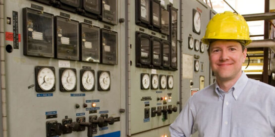 NED-web-pple-024 Een man met een gele helm op en een overhemd met knopen staat voor bedieningspanelen met meters en schakelaars in een industriële omgeving.