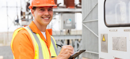 NED-web-pple-026 Een technicus in een oranje veiligheidsuniform en helm staat buiten bij een onderstation, houdt een klembord vast en lacht naar de camera.