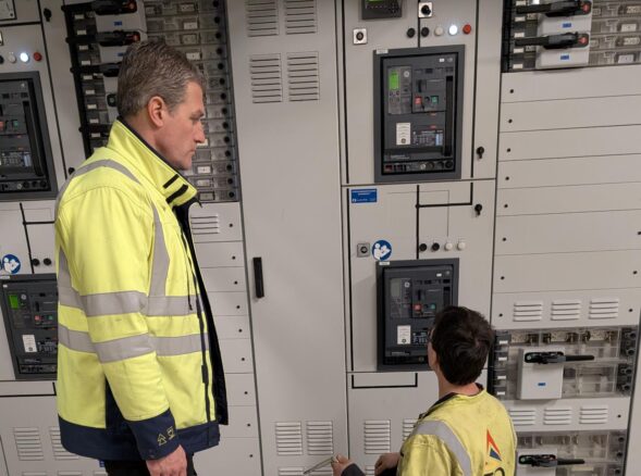 alfred en marci Twee werknemers in veiligheidsvesten inspecteren en werken aan een elektrisch bedieningspaneel in een industriële omgeving.