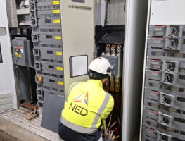 Een arbeider met een veiligheidsvest en helm onderhoudt elektrische panelen met gereedschap dat naast hem op de grond ligt.
